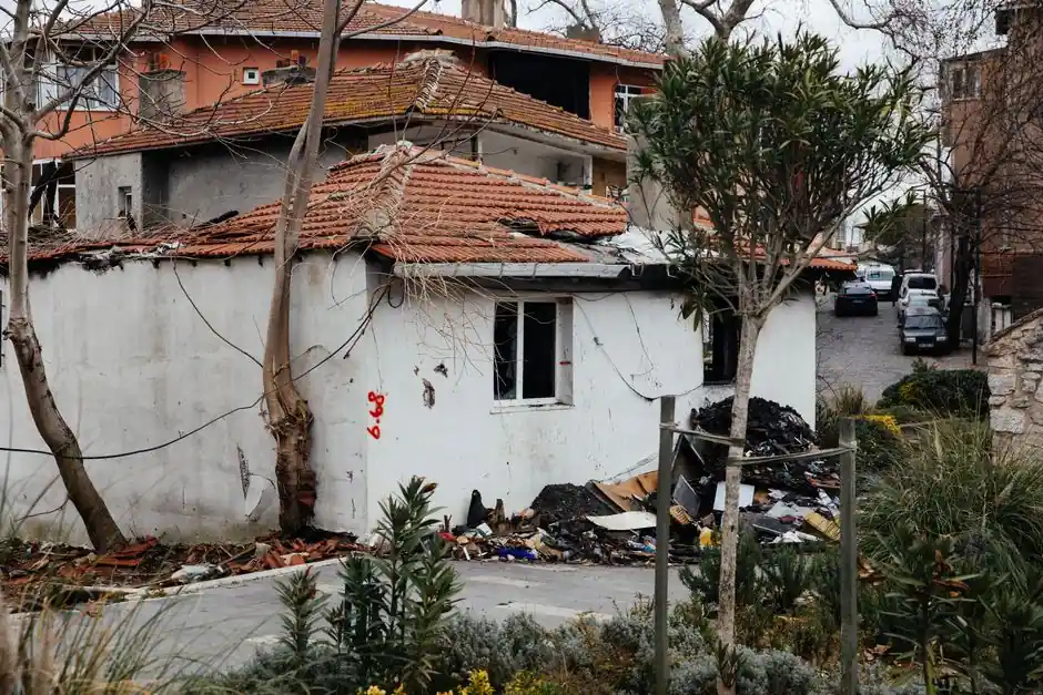 An abandoned house with a damaged roof and debris in an urban neighborhood setting.
