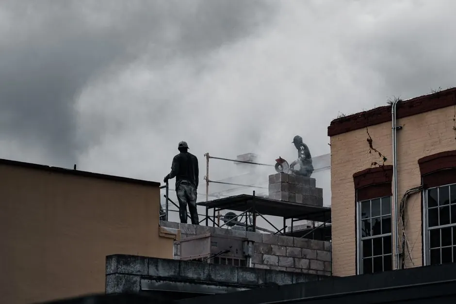 Workers on a multi-storey building site using tools amidst a cloudy backdrop.